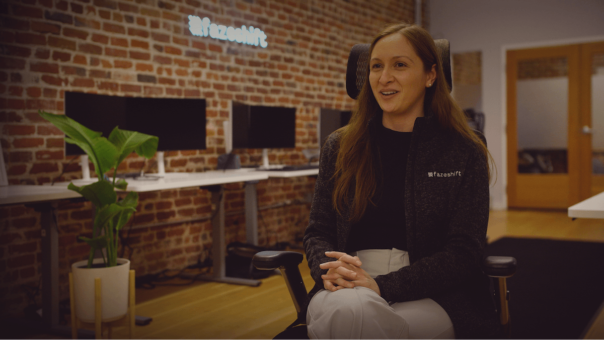 Woman sitting in a modern office with brick walls, wearing a black jacket, in front of computers and a potted plant.